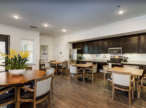 Communal dining room with multiple wooden tables and chairs, a round table with a flower centerpiece, and a full kitchenette with dark cabinets and stainless appliances along one wall.