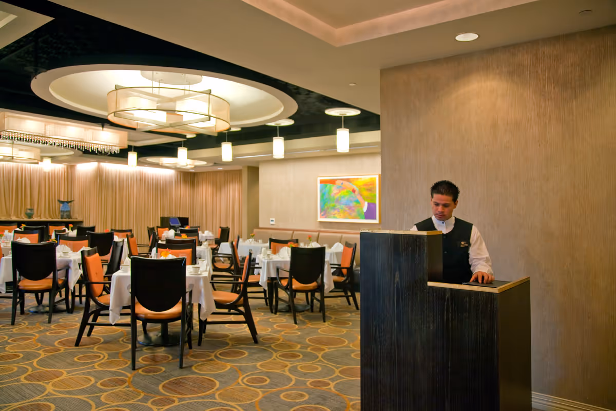 A dining room with multiple tables covered in white tablecloths and set with plates and napkins. Chairs with orange cushions surround the tables. A man in a black vest and white shirt stands behind a dark wooden podium near a beige wall with a colorful abstract painting. The room features modern ceiling lights and a patterned carpet.