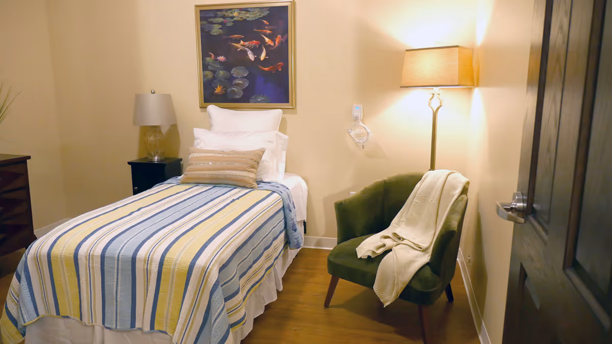 A cozy bedroom with a single bed covered in a striped yellow, blue, and white blanket. There are multiple pillows on the bed. To the right of the bed is a green upholstered chair with a light-colored blanket draped over it, and a tall floor lamp behind it. On the left side of the bed is a nightstand with a table lamp. A framed painting of koi fish and lily pads hangs on the wall above the bed.