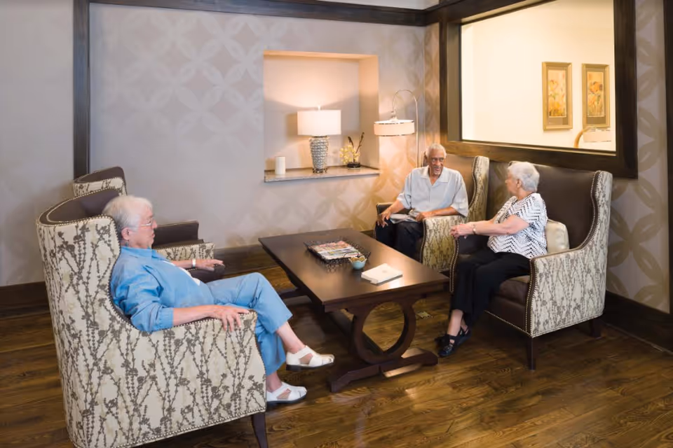 Three elderly individuals sitting and conversing in a cozy living room area with patterned armchairs, a wooden coffee table, a lamp, and decorative wall art.