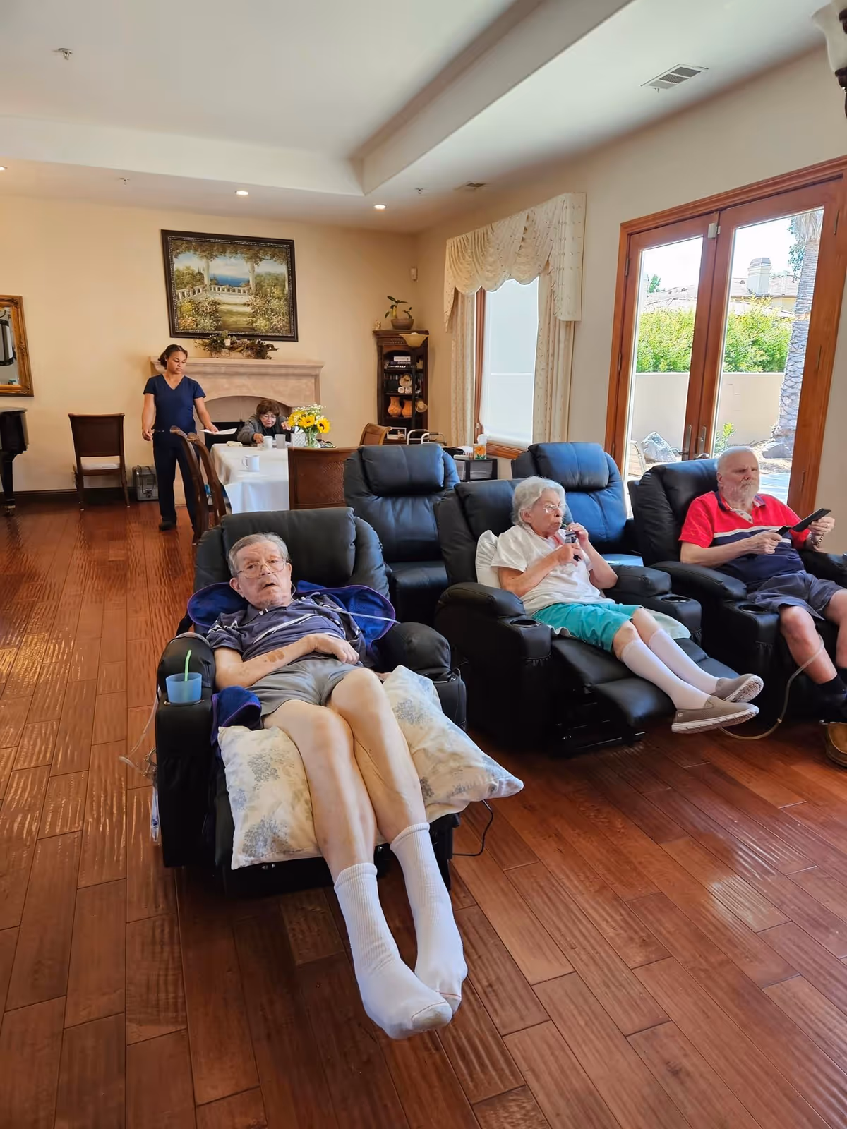 A cozy living room in a senior care facility with three elderly people sitting in black recliner chairs. One elderly man is lying back with a pillow under his legs, an elderly woman is drinking from a cup, and another elderly man is holding a remote control. In the background, a caregiver stands near a dining table where another elderly person is seated. The room has wooden floors, large windows with curtains, and a painting above a fireplace.