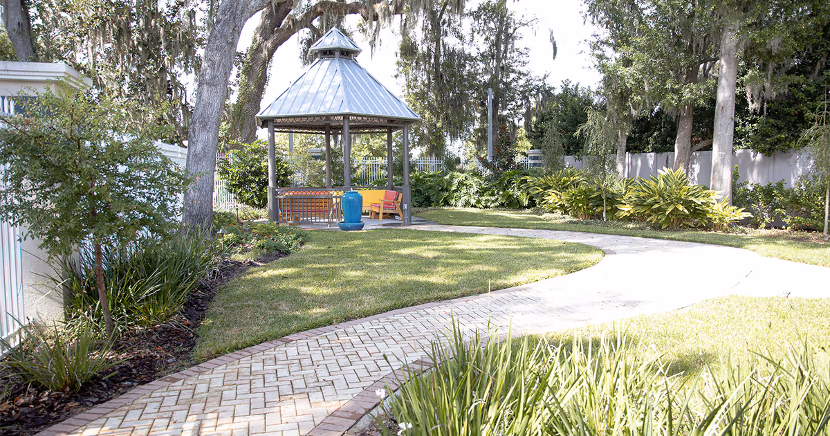 Winding paved path through a landscaped garden leading to a small gazebo surrounded by trees and shrubs.