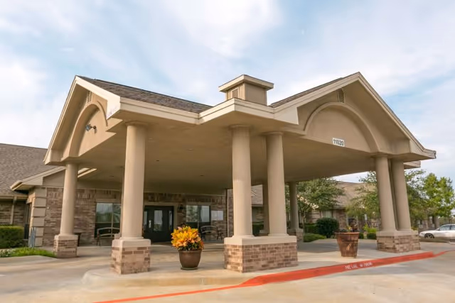 Entrance of a healthcare and rehabilitation facility with a covered drop-off area supported by large columns, a potted plant with yellow flowers in front, and a clear sky above.
