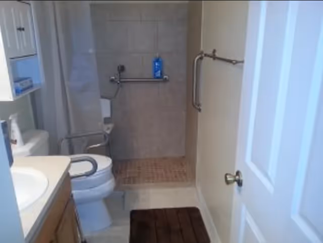 Small accessible bathroom with a toilet, sink, walk-in tiled shower and grab bars.