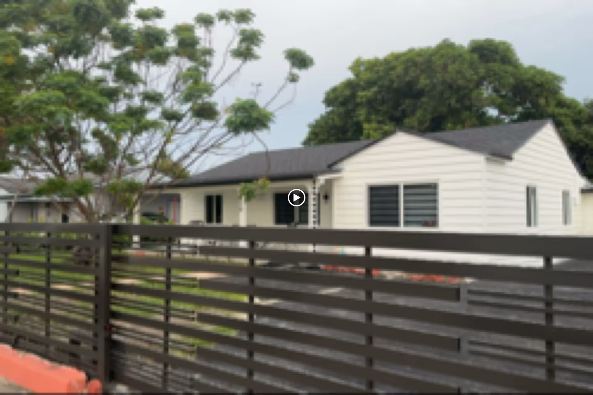 Single-story white residential building behind a dark horizontal fence with trees in the yard.