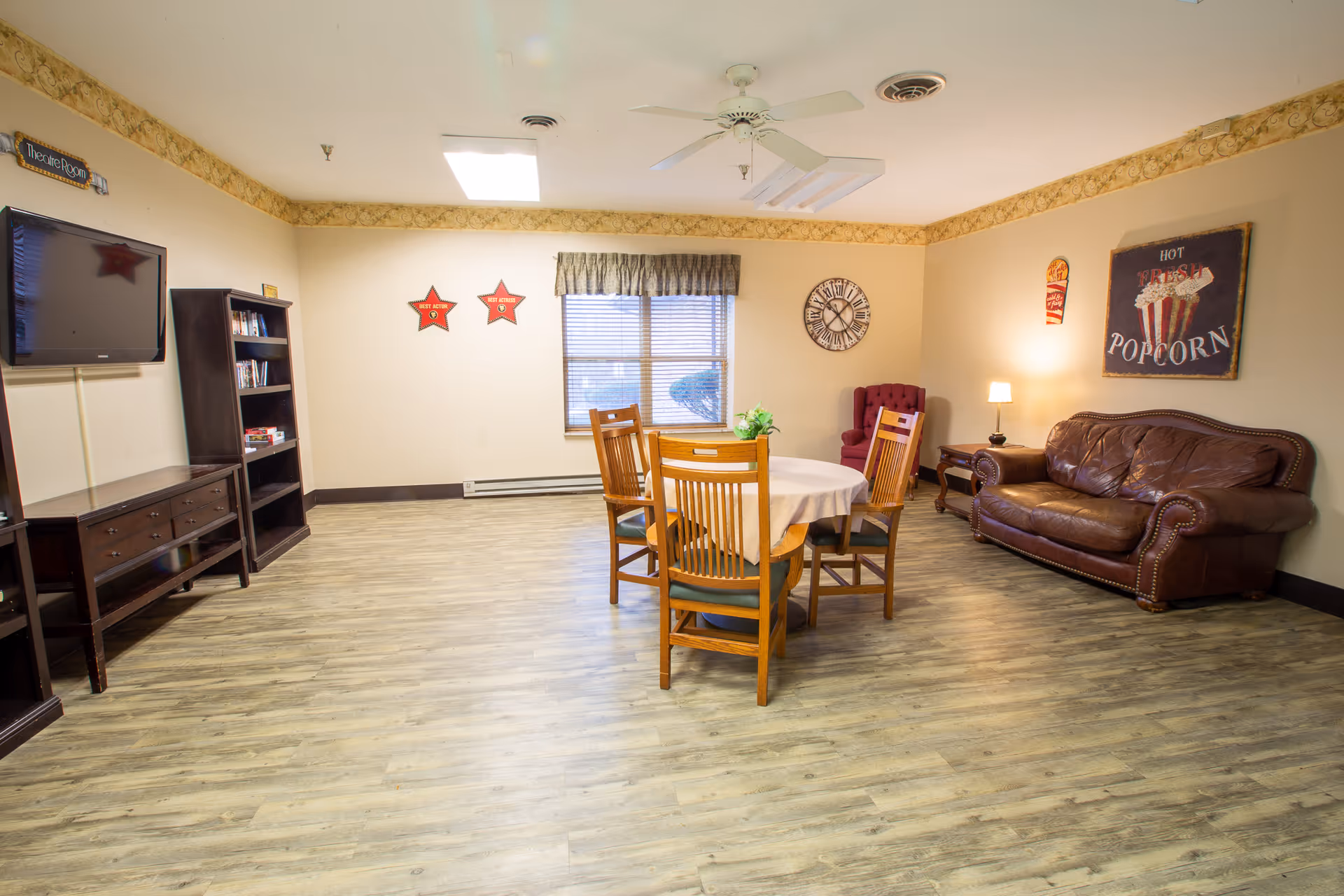 Bright senior living common room with a round table and chairs in the center, a leather sofa and armchair to the right, a TV and shelving on the left, and wood-look flooring.