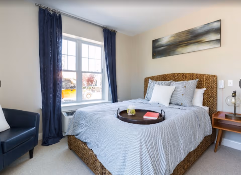 A cozy bedroom with a woven headboard bed covered in a gray and white striped comforter and multiple pillows. A dark wooden tray with a small plant and a red book rests on the bed. To the right of the bed is a wooden nightstand with a modern lamp. A large window with dark blue curtains allows natural light to fill the room. A dark blue armchair with a white pillow is positioned near the window. Above the bed hangs a wide abstract painting in muted tones.