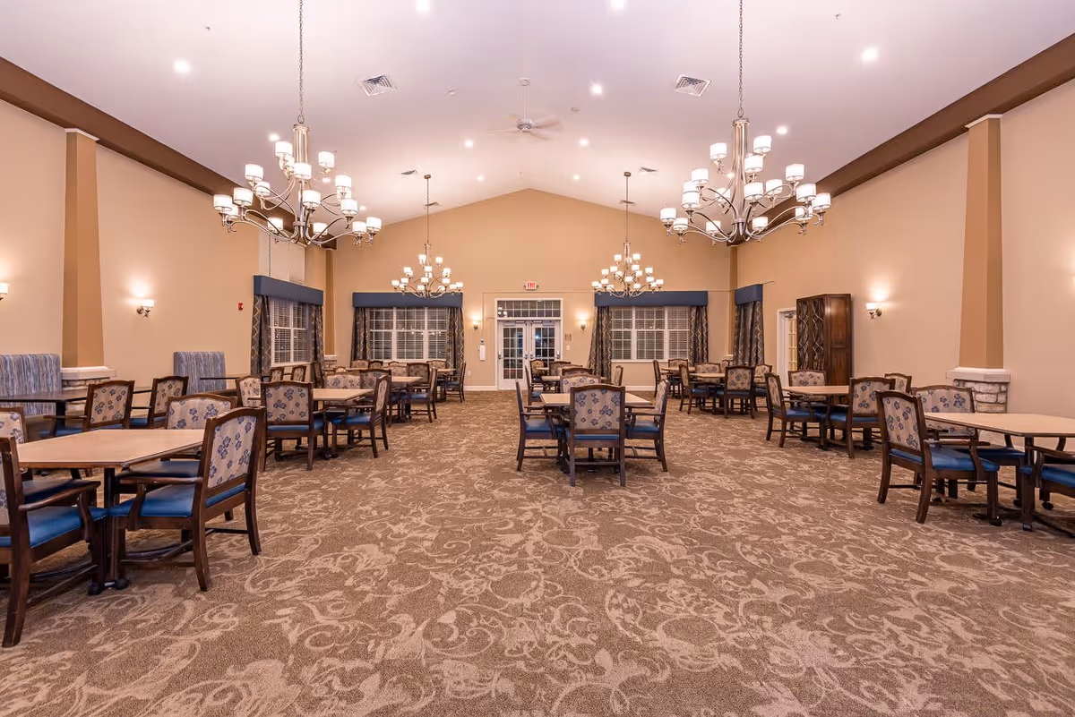 A spacious dining room with multiple wooden tables and chairs arranged neatly. The room features patterned carpet flooring, beige walls, large windows with curtains, and several elegant chandeliers hanging from the ceiling providing warm lighting.