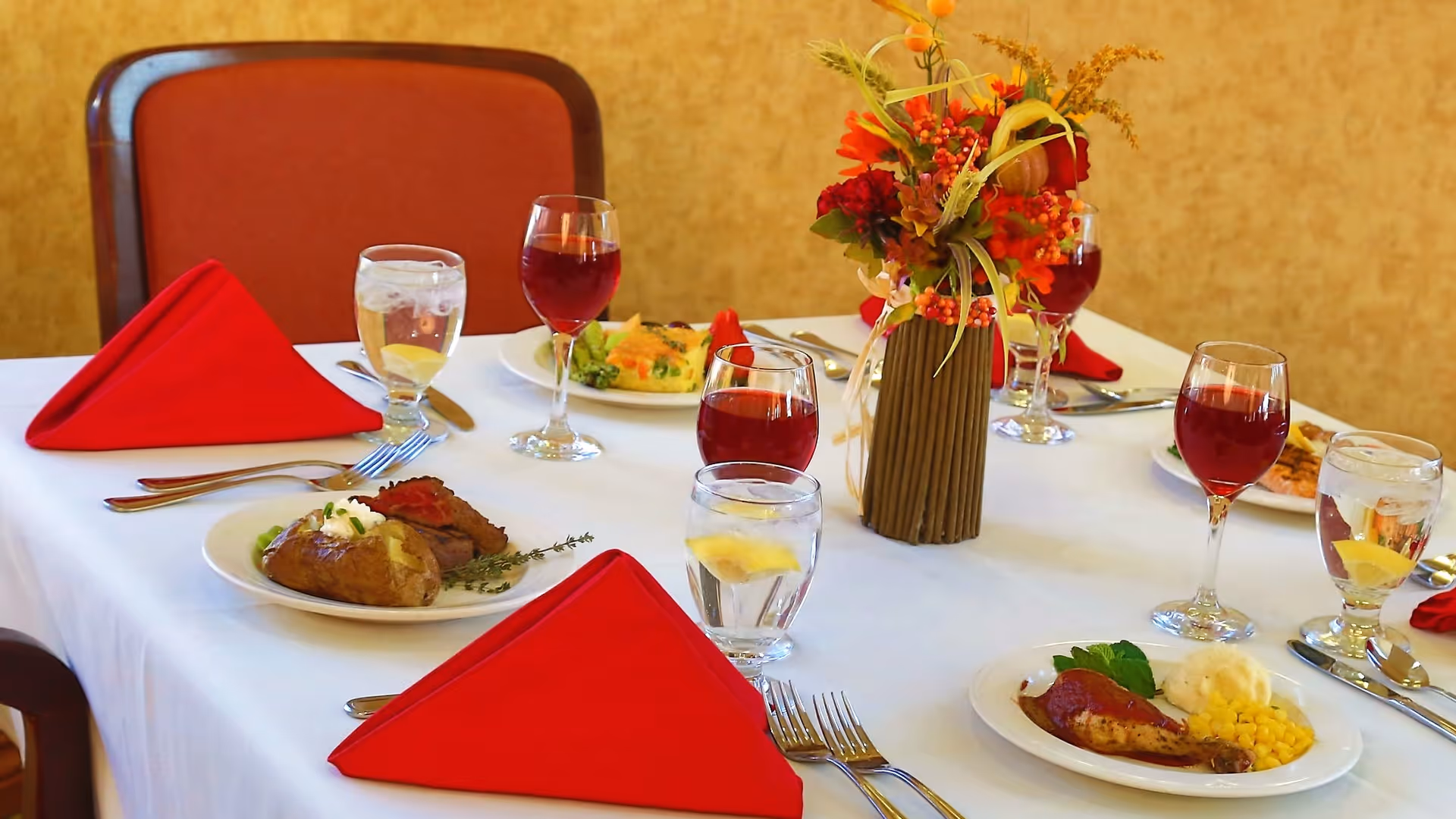 A dining table set for a meal with white tablecloth, red folded napkins, plates of food including baked potato, meat, corn, and mashed potatoes, glasses of water with lemon, glasses of red wine, and a floral centerpiece with autumn-themed flowers and berries.
