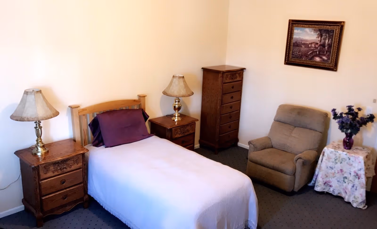 A cozy bedroom with a single bed covered in a white blanket and a purple pillow. There are two wooden nightstands on either side of the bed, each with a brass lamp. A tall wooden dresser stands in the corner. A comfortable beige armchair is next to a small table covered with a floral cloth and a vase of purple flowers. A framed landscape painting hangs on the wall above the armchair.