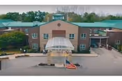 Front exterior view of Mcauley Residence, a multi-story building with a green roof and a covered entrance. An American flag is displayed near the entrance, and there are trees and greenery around the building.
