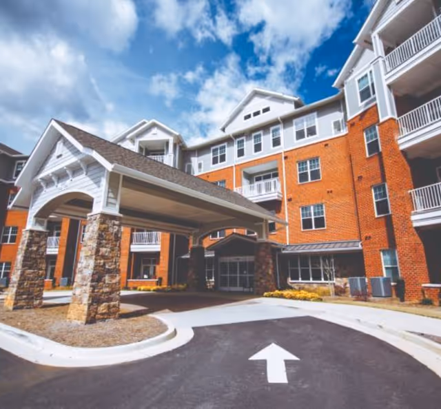 Exterior view of a multi-story senior living facility with a covered entrance supported by stone pillars, red brick walls, white balconies, and a clear blue sky with some clouds.