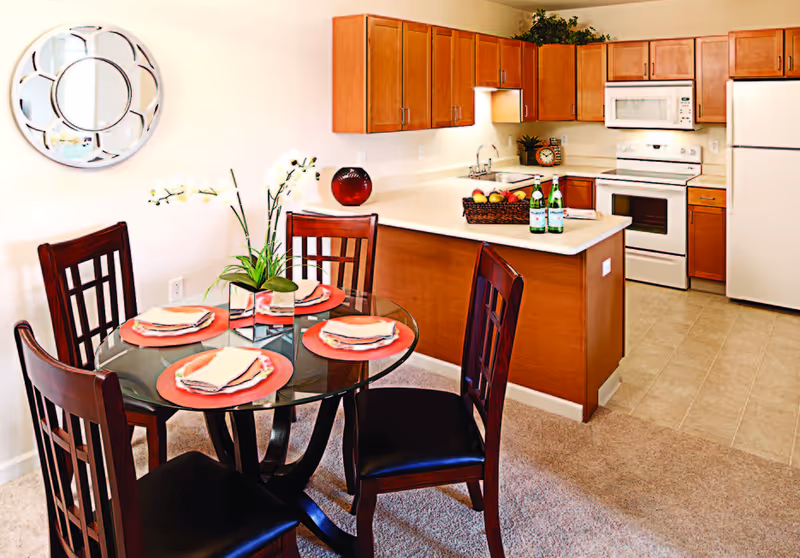 Bright open kitchen with wooden cabinets and a center island and a glass-top dining table set for four in the foreground.