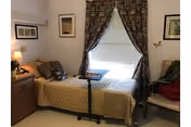 A small, cozy bedroom with a single bed covered in a beige quilt. The bed has several pillows and a small overbed table positioned on top. There is a wooden nightstand with a lamp and some decorative items on the left side of the bed. The window behind the bed is covered with white blinds and floral-patterned curtains. Two framed pictures hang on the walls, and a cushioned chair with a red throw is placed on the right side of the room.