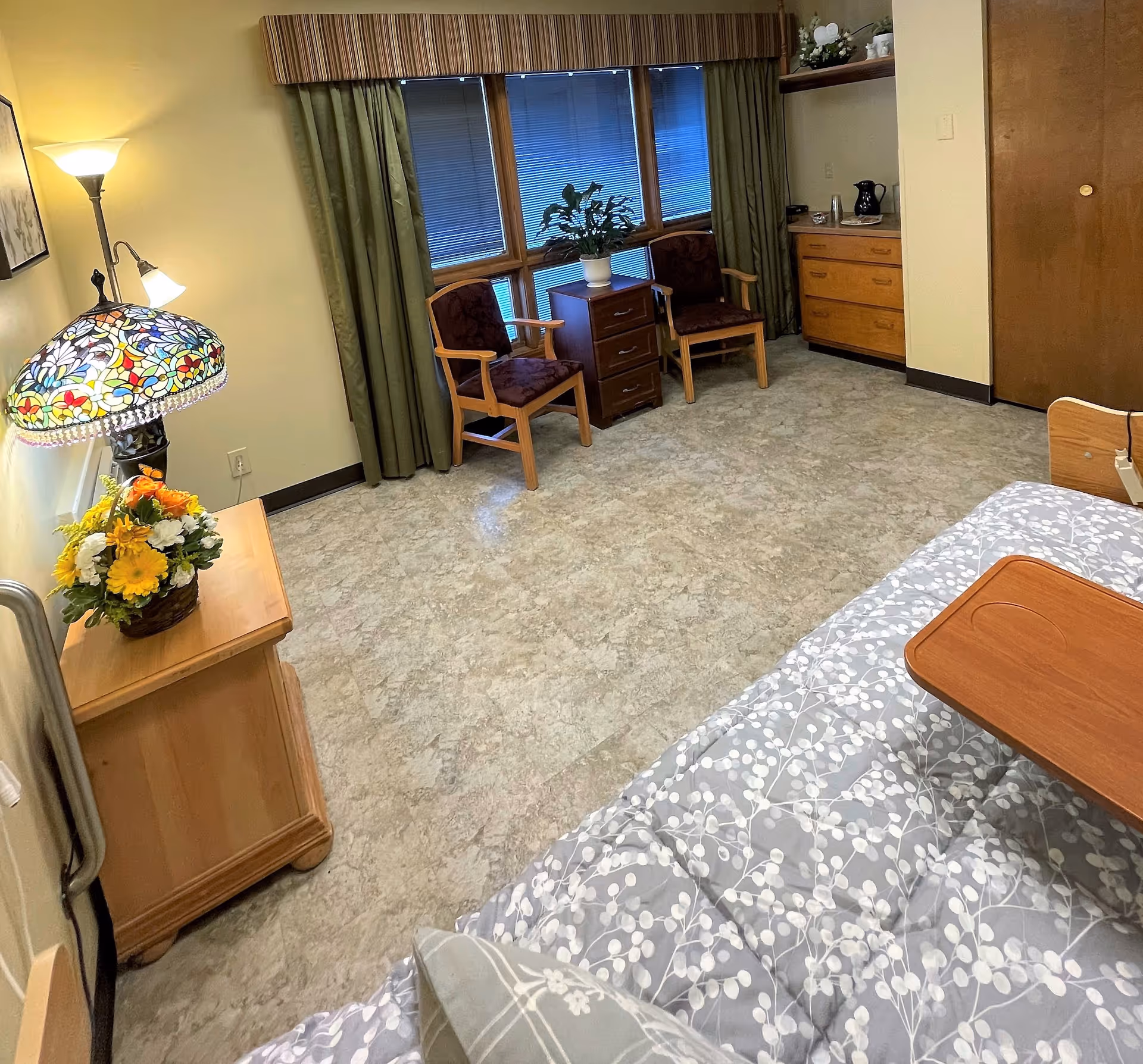 Private senior living bedroom with a bed and overbed tray, a bedside table and lamp, two chairs by a window, and a dresser.