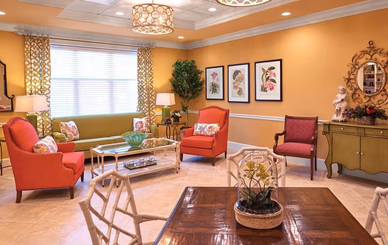 A cozy living room with warm yellow walls, featuring a green sofa with floral pillows, two red armchairs, a glass coffee table, and a wooden dining table with white chairs. The room is decorated with framed botanical prints, a potted plant, a decorative mirror, and a small statue on a green sideboard. The space is well-lit with natural light from a large window and ceiling lights.
