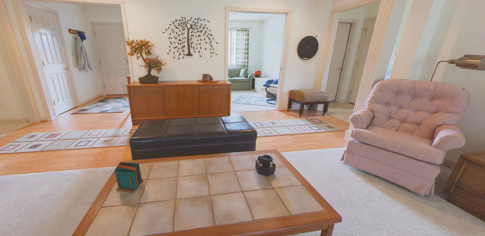A cozy living room area with a pink upholstered armchair, a tiled coffee table with small decorative items, and a black leather bench. In the background, there is a wooden cabinet with a potted plant and wall art above it. Multiple doorways lead to other rooms, one of which has a green armchair and a window with curtains.