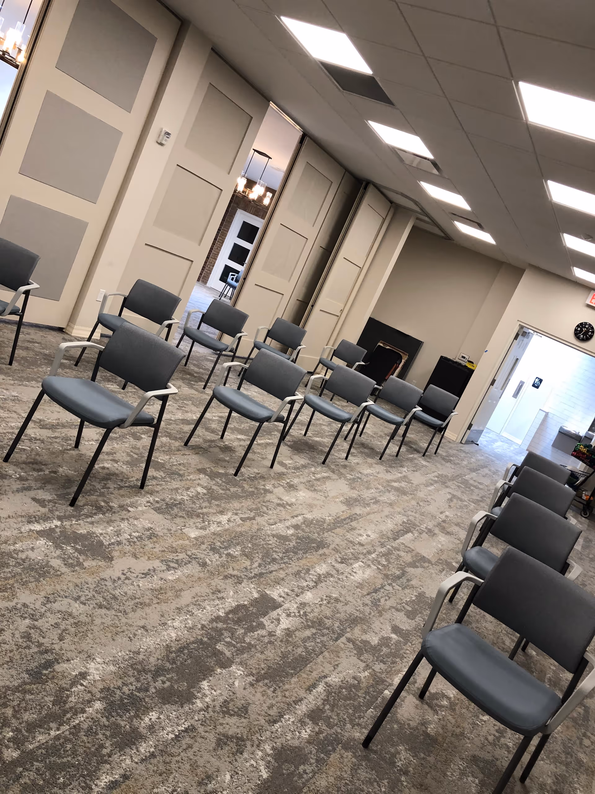 A room with multiple rows of gray chairs arranged in a spaced-out manner on a patterned carpet floor. The room has beige walls with paneling, a drop ceiling with fluorescent lights, and an open doorway leading to another area with a clock above it.