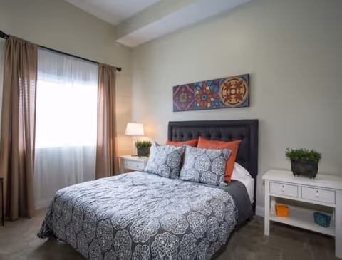 A cozy bedroom with a large bed featuring a dark gray tufted headboard, patterned gray and white bedding, and orange accent pillows. There are two white nightstands on either side of the bed, each with a small green plant. A lamp with a white shade is on the left nightstand. The room has beige walls, a window with sheer white curtains and brown drapes, and a colorful abstract painting above the headboard.