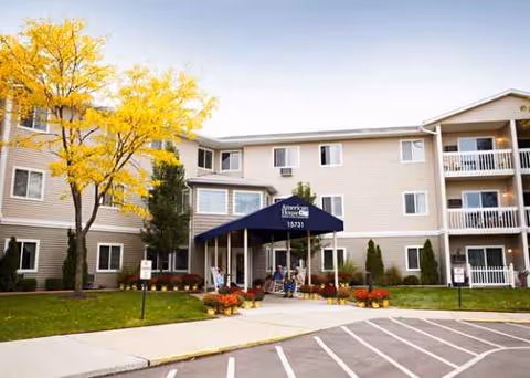 Beige senior living building with blue entrance canopy and autumn trees.