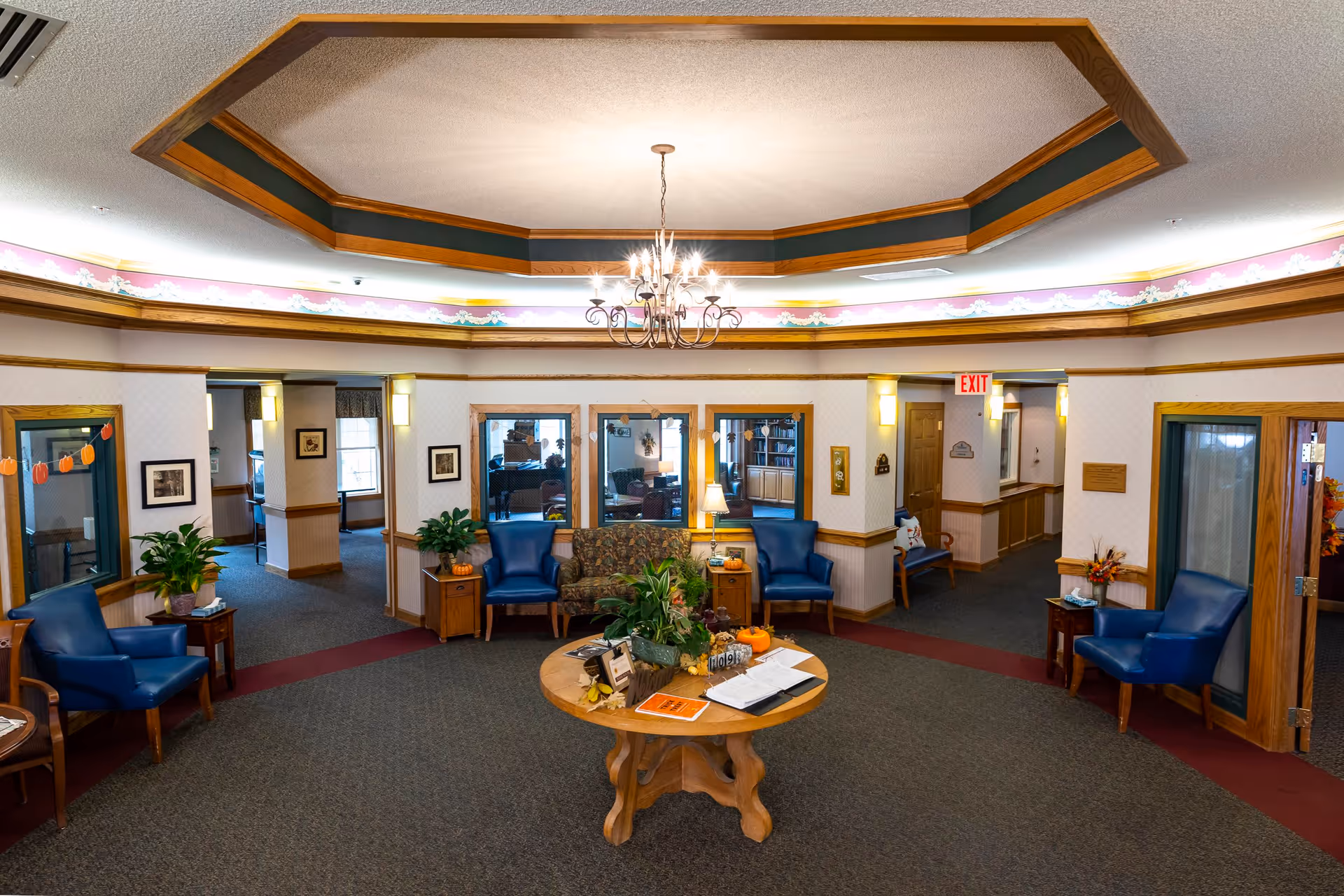 A spacious and well-lit common area in an assisted living facility featuring a round wooden table with plants and decorations in the center. Surrounding the table are several blue armchairs and a floral-patterned sofa. The ceiling has a decorative wooden trim with a chandelier hanging in the middle. The walls are adorned with framed pictures and plants, and there are multiple doorways leading to other rooms.