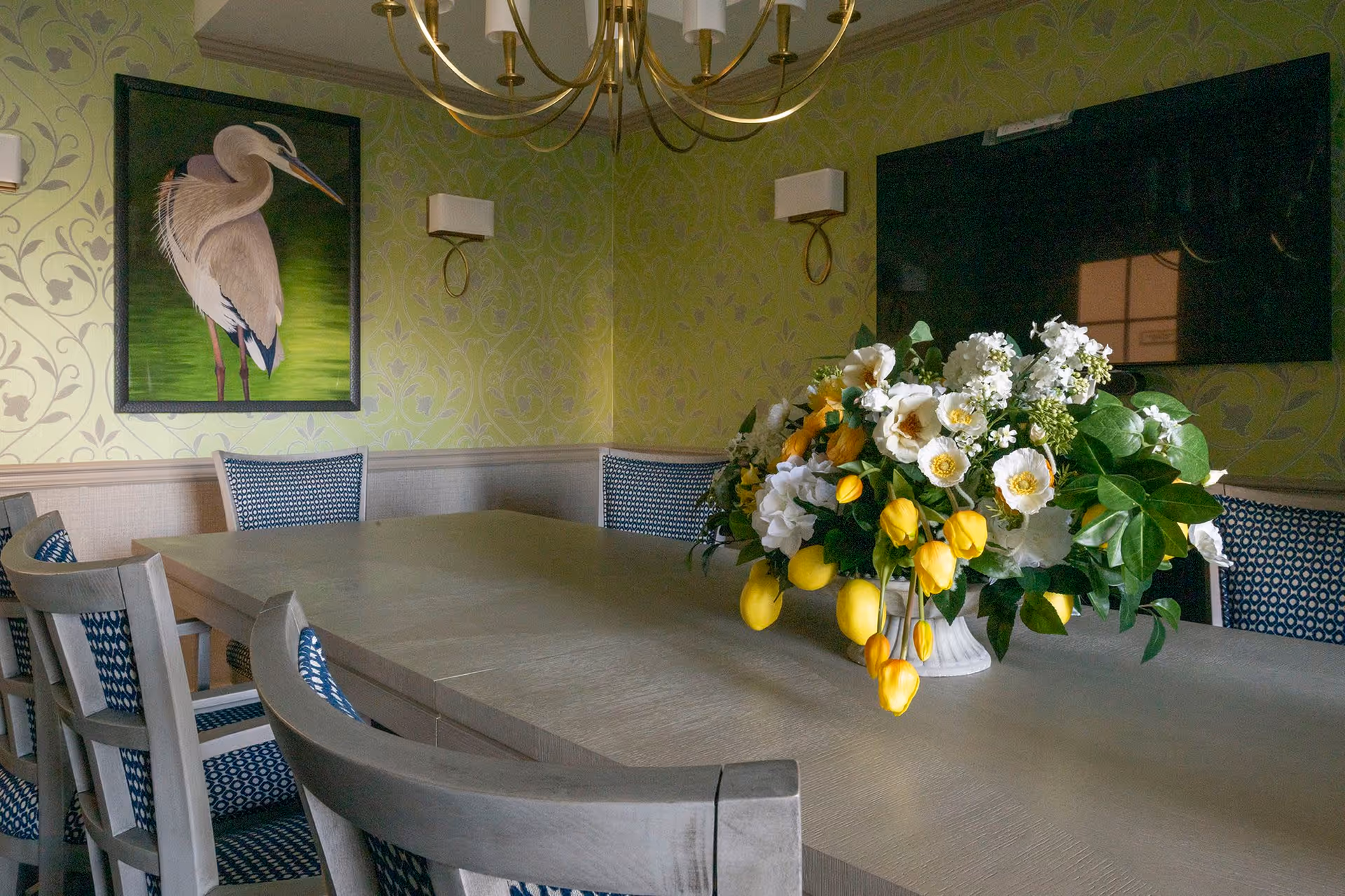 A dining room with a long wooden table surrounded by chairs with blue patterned cushions. A large floral arrangement with white and yellow flowers sits in the center of the table. The walls are decorated with green patterned wallpaper, a painting of a bird, two wall sconces, and a large flat-screen TV.