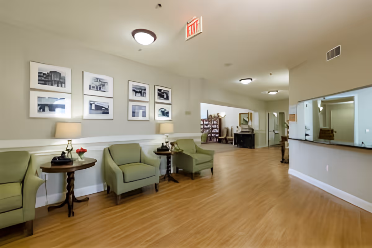 A spacious interior common area with light green armchairs and small wooden tables with lamps and flowers. The walls are decorated with framed black and white photographs. The floor is wooden, and the ceiling has round light fixtures. There is a reception window on the right side and an exit sign on the ceiling.