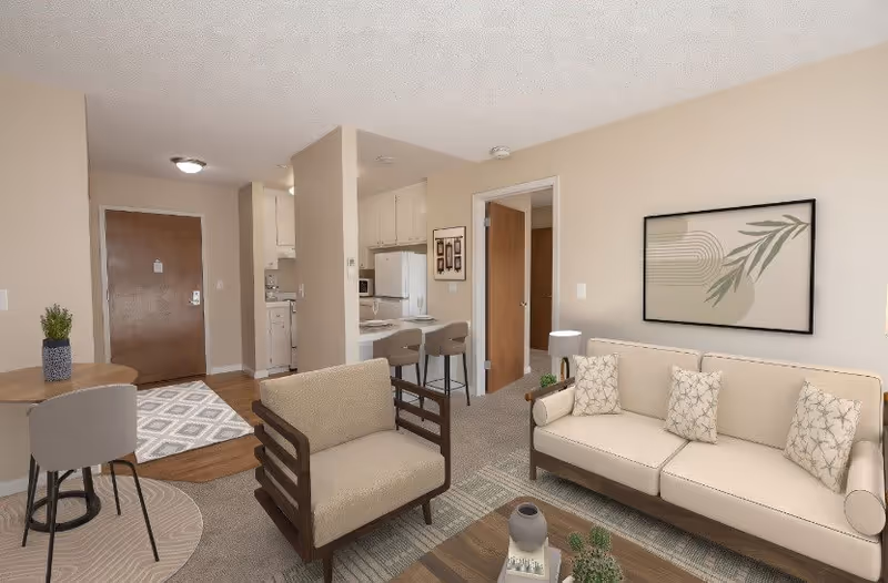 A cozy senior living apartment interior featuring a beige sofa with patterned cushions, a matching armchair, a wooden coffee table with decorative items, a small dining table with two chairs, and a compact kitchen area with white cabinets and appliances. The walls are painted light beige with framed artwork, and the entrance door is visible in the background.