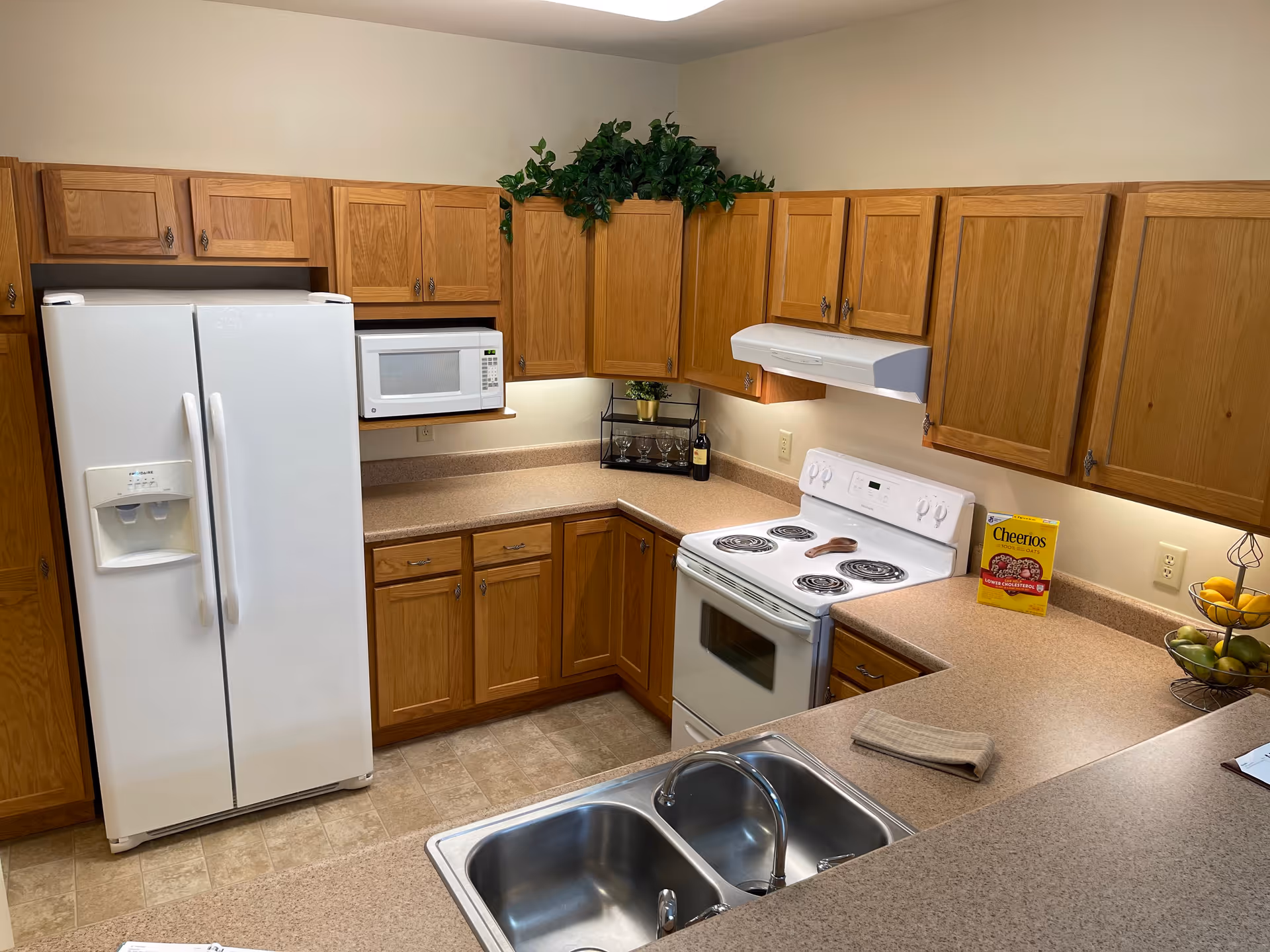 A kitchen with wooden cabinets, a white refrigerator with water dispenser, a white microwave, a white electric stove with four coil burners, and a double stainless steel sink. The countertop is beige with a box of Cheerios cereal, a fruit basket with lemons and green apples, a small towel, and some decorative greenery on top of the cabinets.