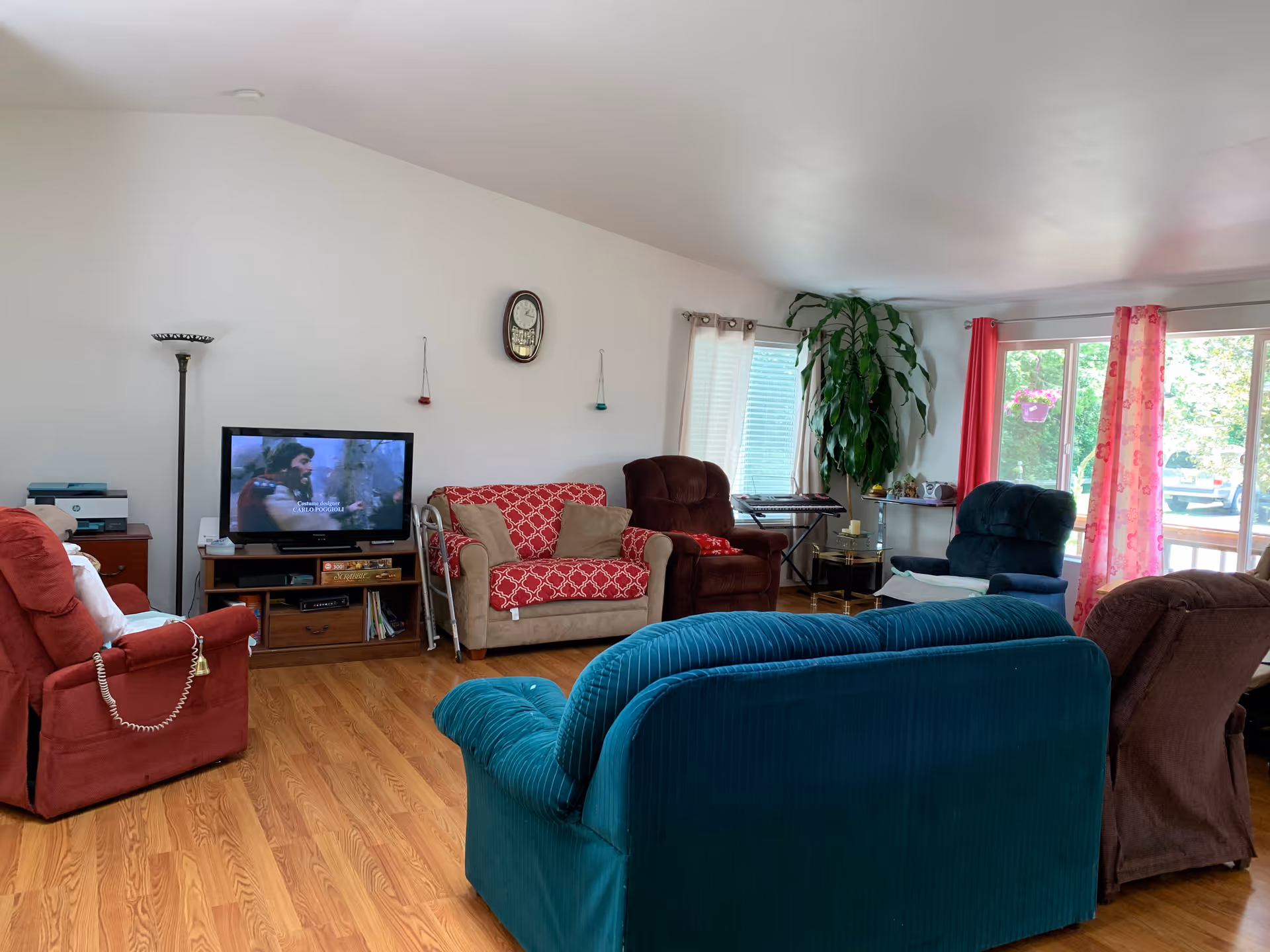 A cozy living room with multiple upholstered chairs and sofas arranged around a television on a wooden stand. The room has hardwood floors, a tall floor lamp, a wall clock, and large windows with red and pink curtains letting in natural light. There is a large green plant near the window and a keyboard on a stand in the corner.