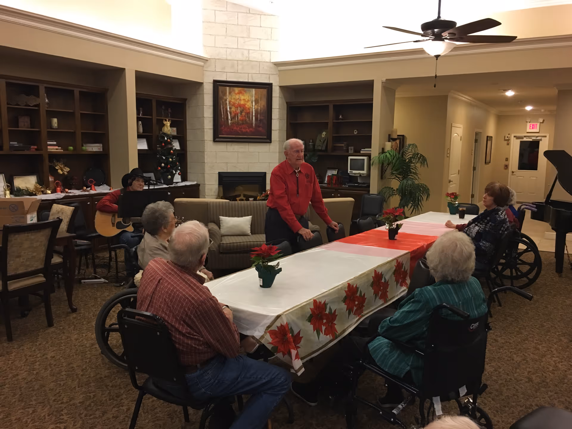 A group of elderly people, some in wheelchairs, gathered around a long table decorated with a festive tablecloth and poinsettia plants in a cozy living room area. A man in a red shirt is standing and speaking to the group, while another person is playing a guitar in the background near bookshelves and a fireplace. The room has warm lighting, a ceiling fan, and a piano in the corner.
