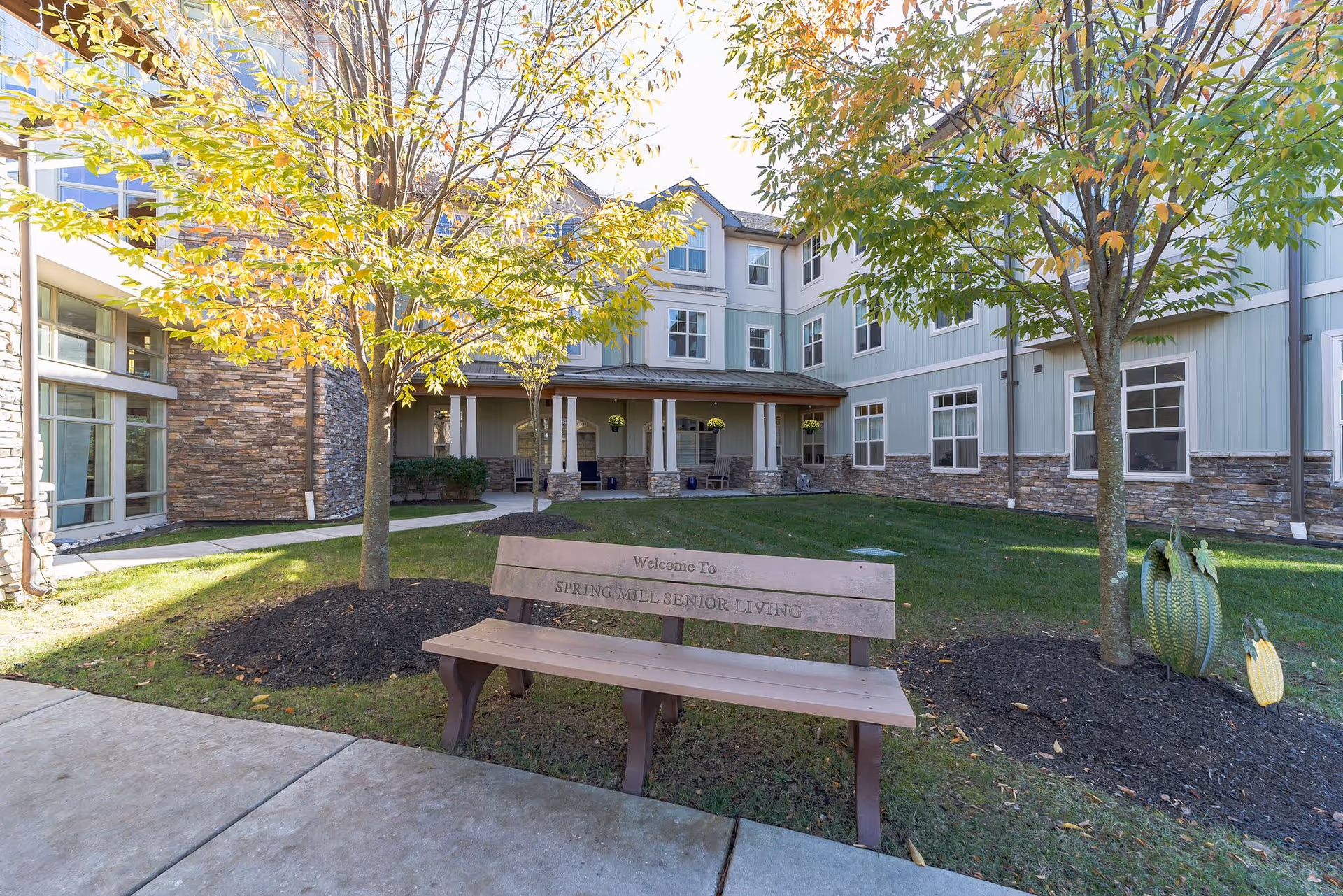 Outdoor courtyard area of Spring Mill Senior Living facility with a wooden bench that has 'Welcome To Spring Mill Senior Living' engraved on it. The courtyard features green grass, two trees with autumn leaves, a concrete walkway, and a building with stone and light blue siding in the background.