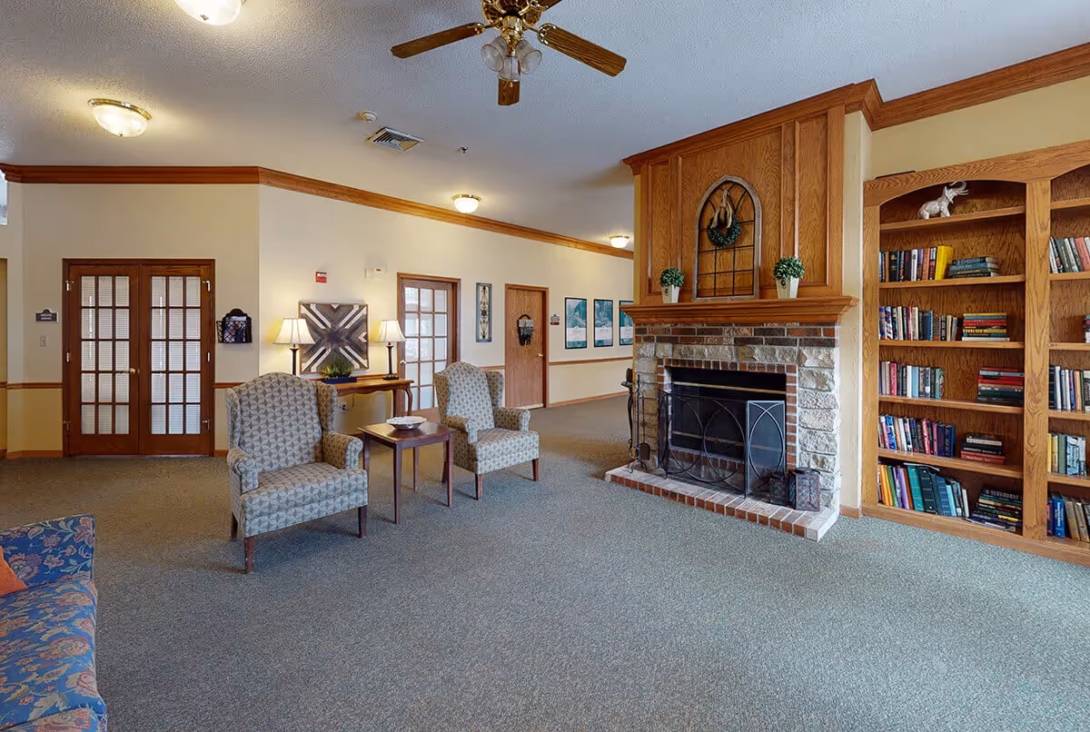 A cozy living room area with two patterned armchairs and a small wooden table between them. There is a stone fireplace with a wooden mantel decorated with two small potted plants and a decorative window frame above it. To the right of the fireplace is a built-in wooden bookshelf filled with books and decorative items. The room has beige walls with wooden trim, carpeted flooring, and a ceiling fan with wooden blades. There are also two lamps on a console table against the wall and several framed pictures hanging on the walls.