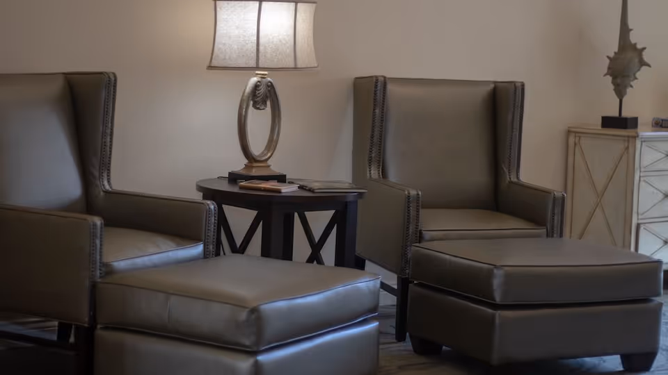 Two gray leather armchairs with matching ottomans are positioned around a small round wooden side table with a decorative lamp and some books on it. In the background, there is a light-colored cabinet with a decorative sculpture on top. The setting appears to be a cozy and well-lit living area.