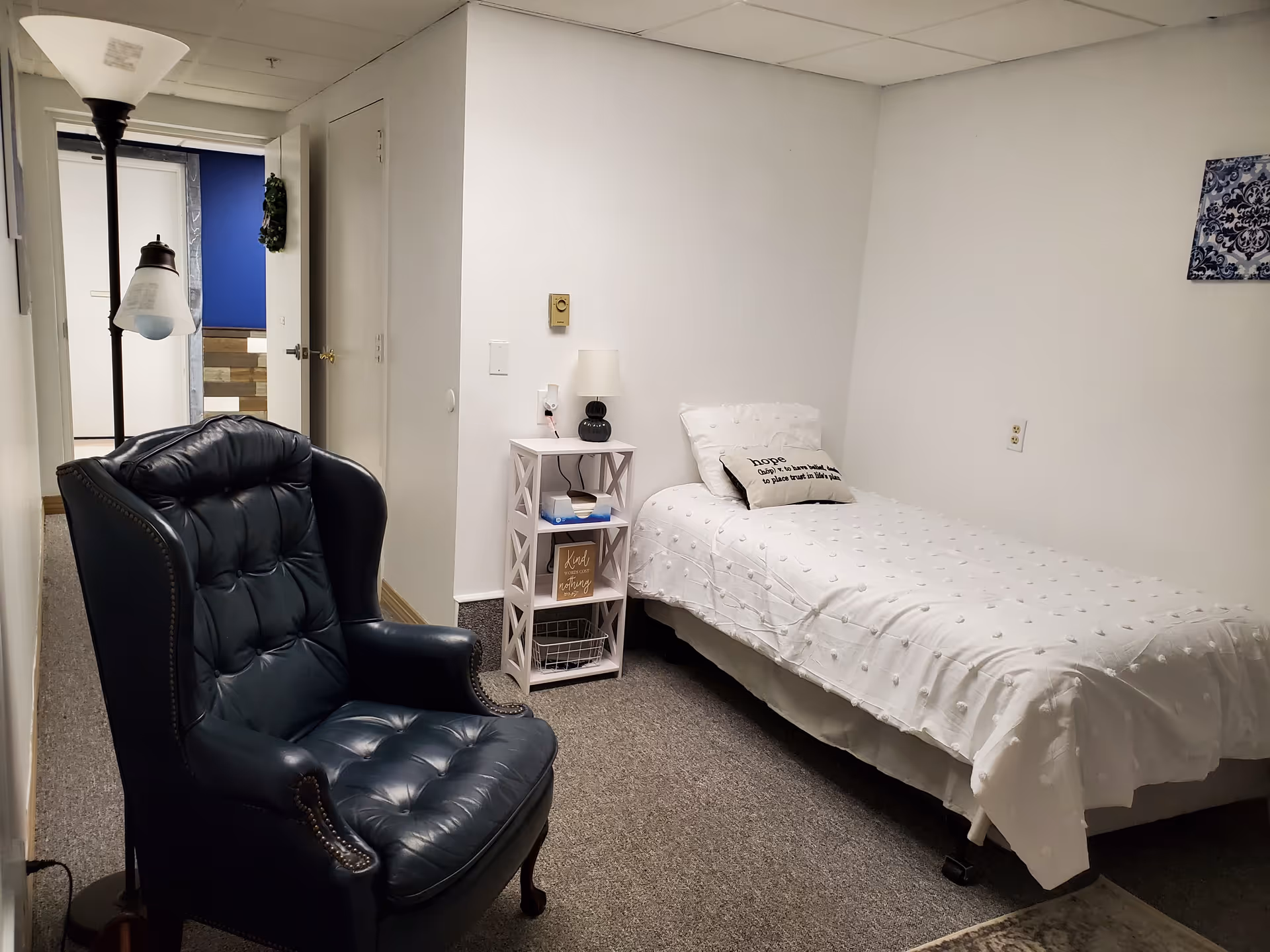 Small single-occupancy bedroom with a twin bed, bedside table and lamp, and a dark upholstered armchair.