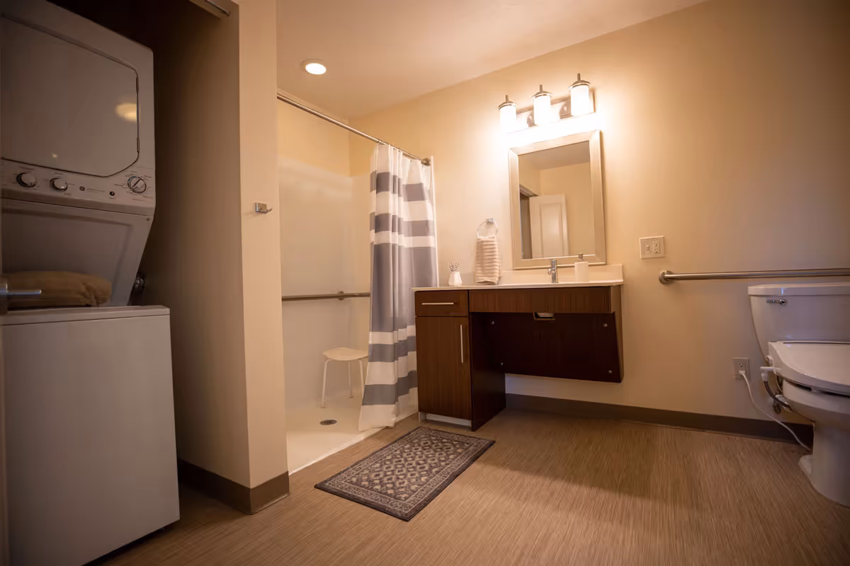 Accessible bathroom with a walk-in shower and stool, striped shower curtain, vanity with mirror and lighting, toilet, and a stacked washer-dryer.