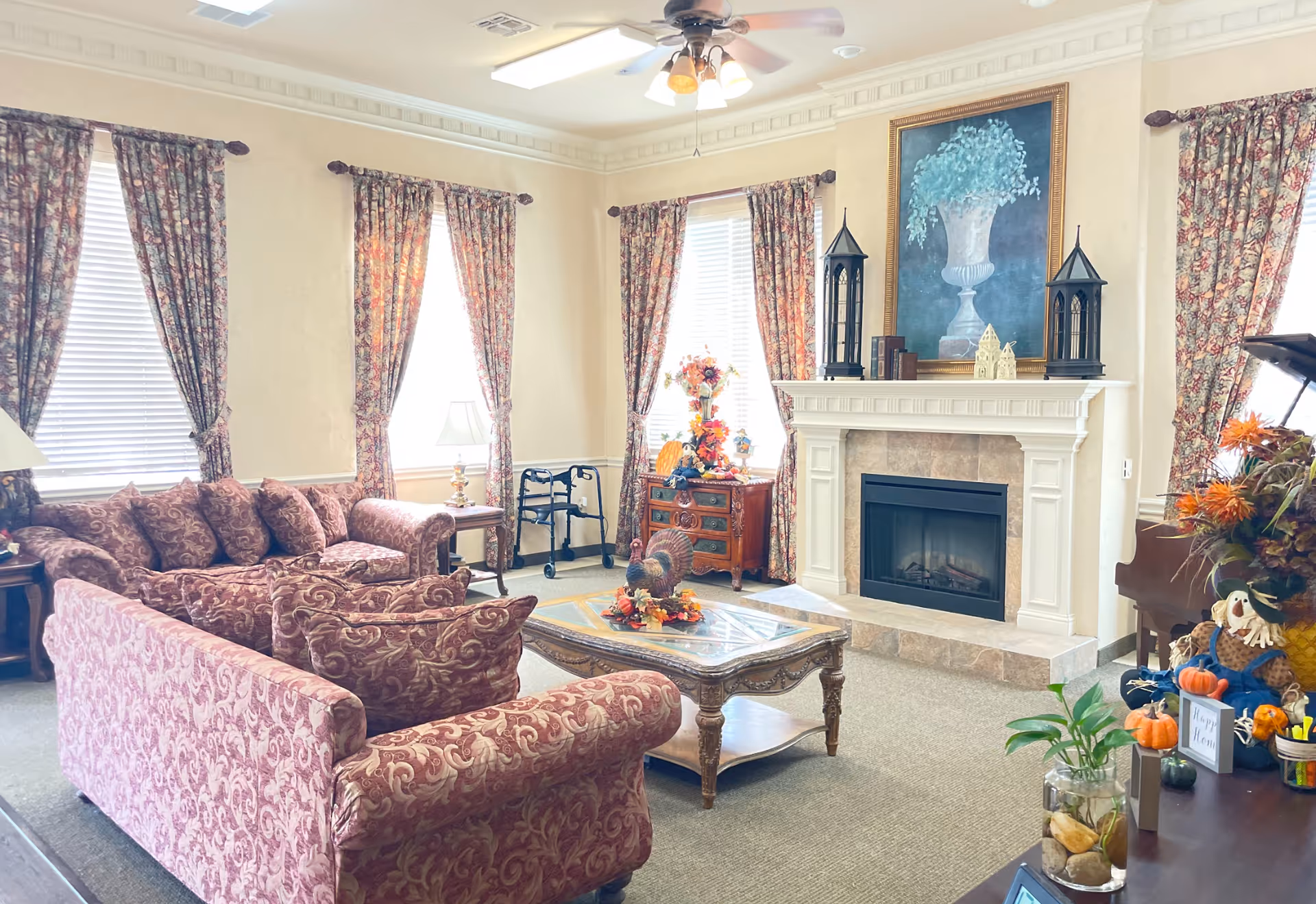 A cozy living room in Colonial Lodge Assisted Living featuring patterned maroon sofas and armchairs arranged around a wooden coffee table with decorative items. The room has three windows with floral curtains, a white fireplace with a large framed painting above it, and various autumn-themed decorations including pumpkins and flowers. A walker is positioned near a side table, and a piano is partially visible on the right side.