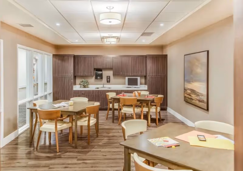 A well-lit room with several wooden tables and chairs arranged for dining or activities. The back wall features a kitchenette with dark wood cabinets, a sink, a microwave, and a small TV. There is a large framed painting on the right wall and large windows on the left side letting in natural light.