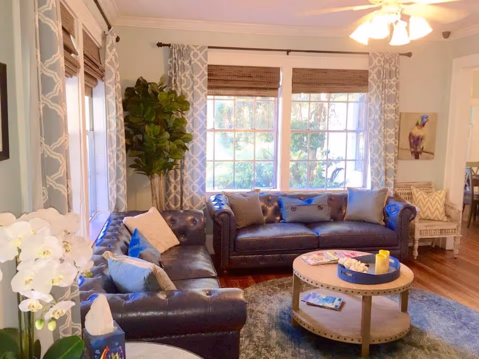 A cozy living room with two dark leather sofas adorned with various throw pillows. A round wooden coffee table with magazines and a candle sits on a blue patterned rug in the center. Large windows with patterned curtains and bamboo blinds let in natural light. A tall green plant is in the corner, and a ceiling fan with lights is above. There is a wicker chair with cushions and a colorful bird painting on the wall.