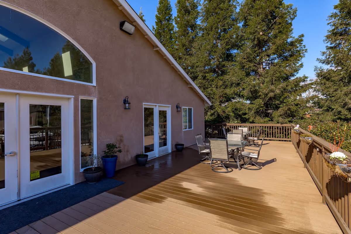 Outdoor patio area at Shearwater Senior Living with wooden deck flooring, several chairs and tables, potted plants, and a railing with flower pots. The building exterior is beige with large windows and doors, surrounded by tall green trees under a clear blue sky.