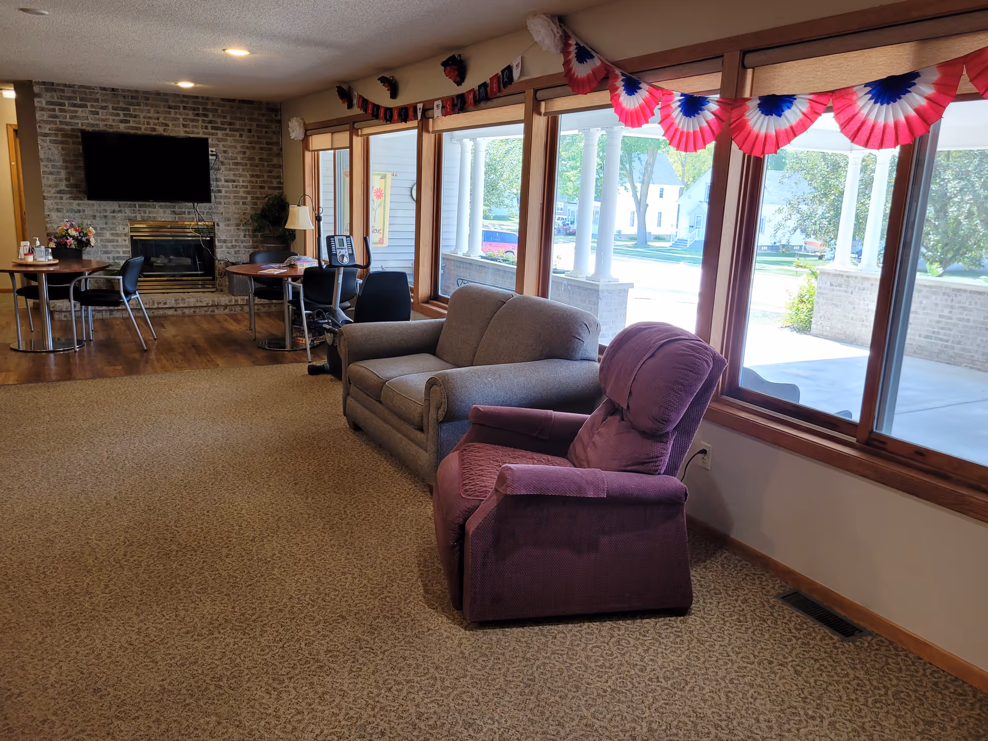 A cozy living room area with a purple recliner and a gray loveseat near large windows decorated with red, white, and blue bunting. In the background, there are two small round tables with chairs, a brick fireplace with a mounted TV above it, and an exercise bike. The room has carpeted flooring and wood trim around the windows and doorways.