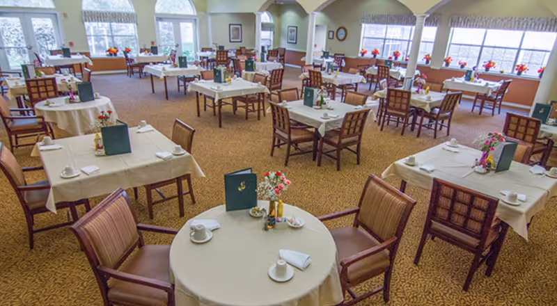 A spacious dining room with multiple tables covered in white tablecloths, each set with cups, napkins, menus, and flower centerpieces. The room has large windows allowing natural light to fill the space, and wooden chairs are arranged around the tables.