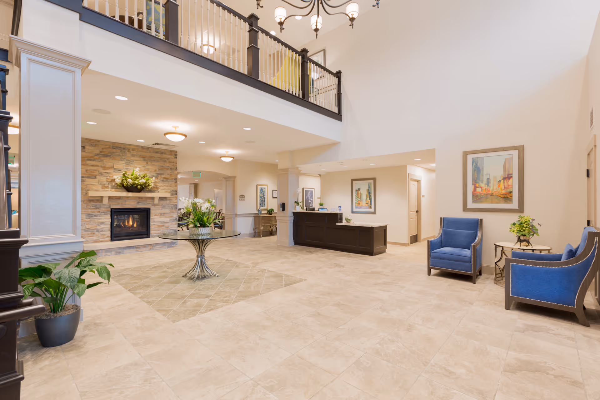 Spacious senior living facility lobby with beige tiled floors, a stone fireplace with a floral arrangement on the mantel, a round glass table with plants in the center, a dark wood reception desk, blue upholstered chairs with side tables, framed artwork on the walls, and a second-floor balcony railing above.