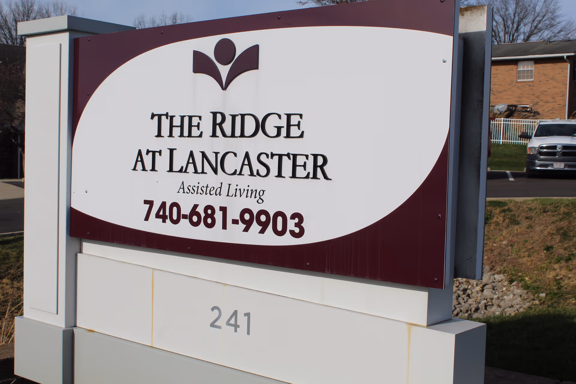 Outdoor sign for The Ridge at Lancaster Assisted Living with contact phone number 740-681-9903 and address number 241, with a parking lot and building visible in the background.