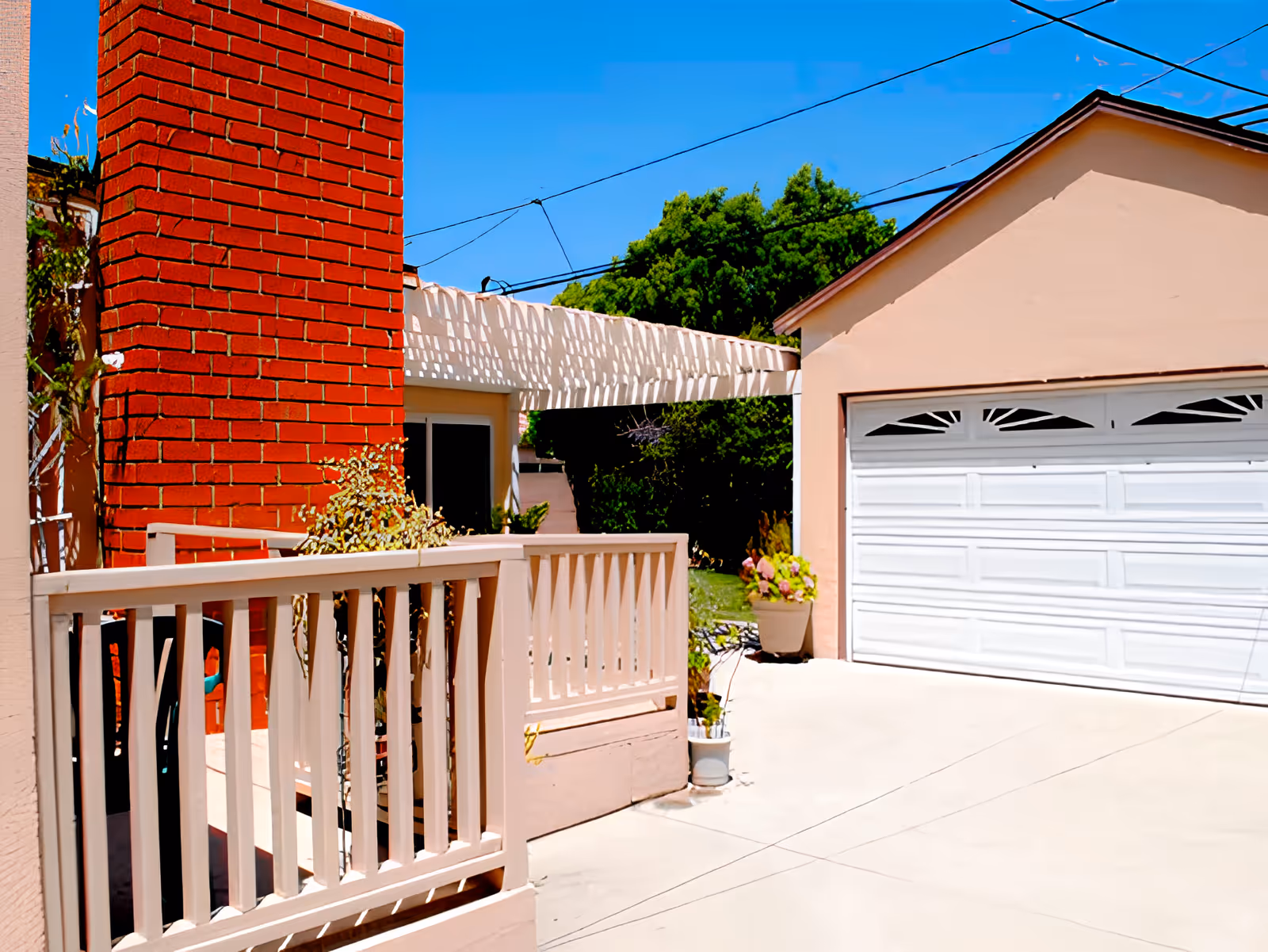 Outdoor view of a residential facility showing a beige building with a white garage door, a red brick chimney, a white wooden railing, potted plants, and a clear blue sky with overhead utility wires.