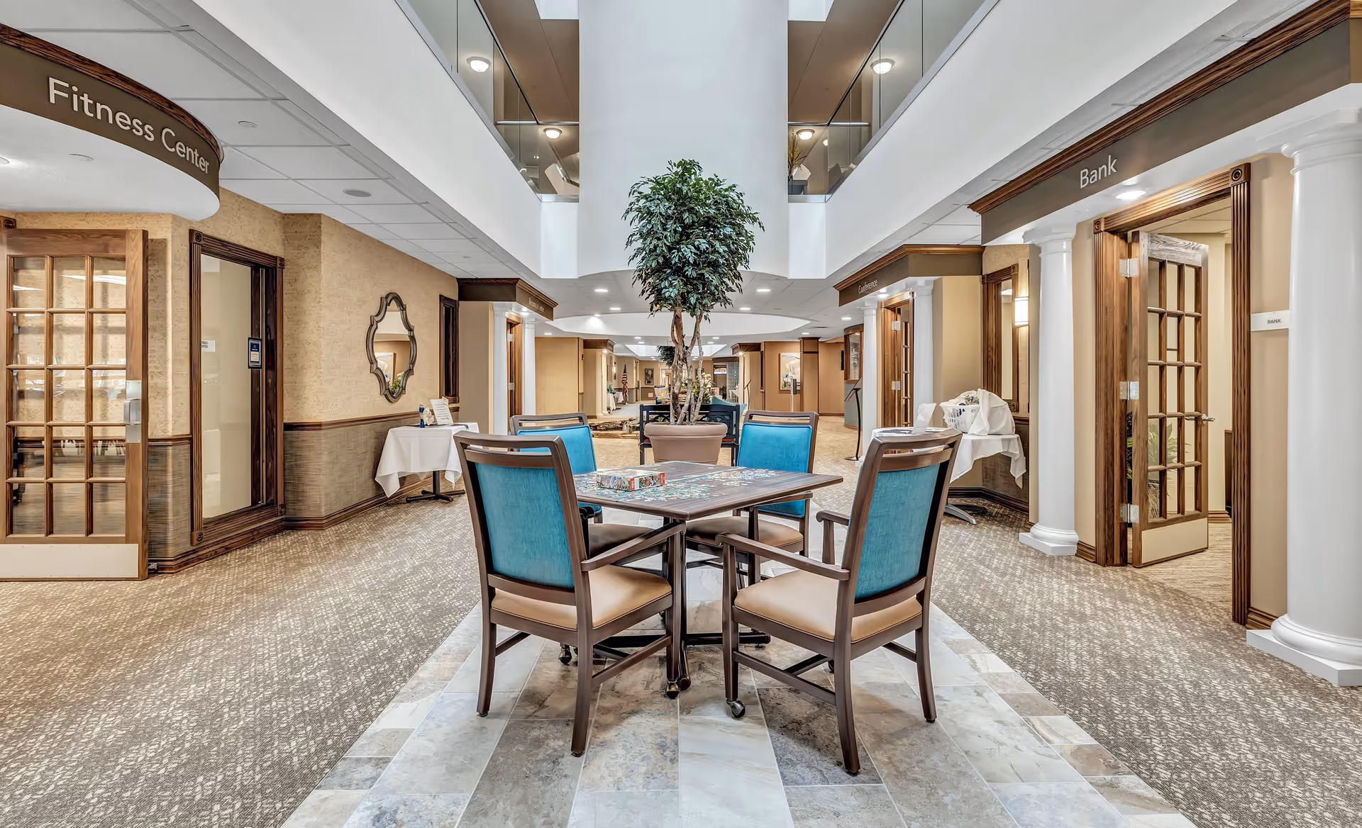 Interior view of a retirement community hallway with a central seating area featuring a table and four chairs with blue backs. The hallway has doors labeled Fitness Center, Bank, and Conference, with beige walls, carpeted floors, and a potted plant in the middle. The ceiling has recessed lighting and a second-floor balcony overlooking the area.