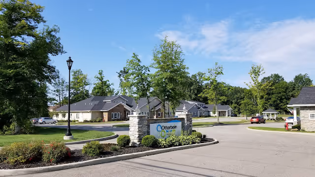Entrance drive and landscaped roundabout with an 'Otterbein' sign and single-story senior living buildings under a blue sky.