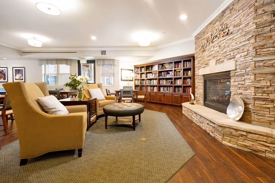 A warm lounge with upholstered armchairs, a round ottoman, built-in bookshelves and a stone fireplace.