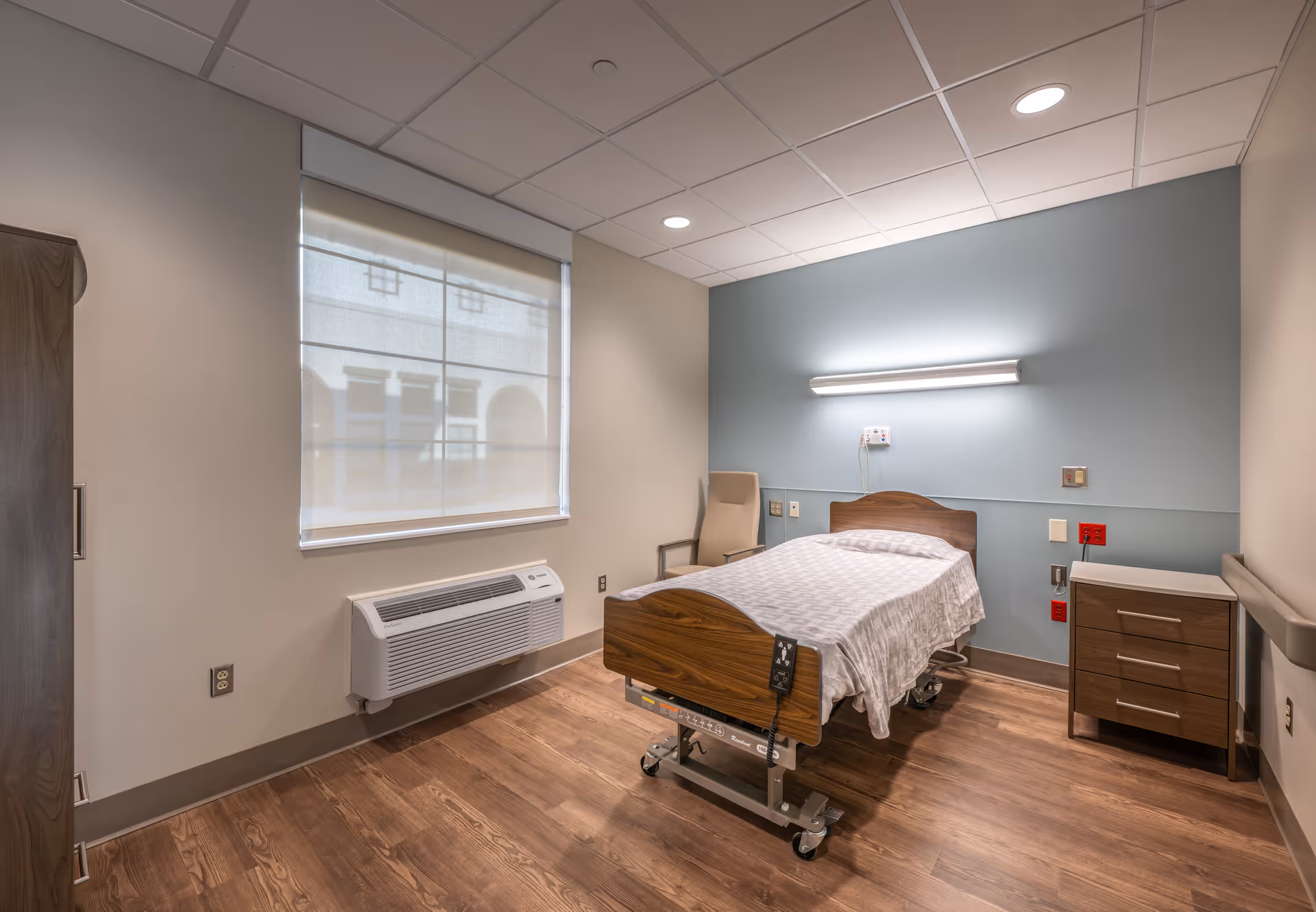 A simple, clean patient or resident room with a single hospital-style bed, a wooden nightstand with drawers, a beige chair, a window with a white roller shade, and a wall-mounted light above the bed. The room has wood-look flooring and light-colored walls with a blue accent wall behind the bed.
