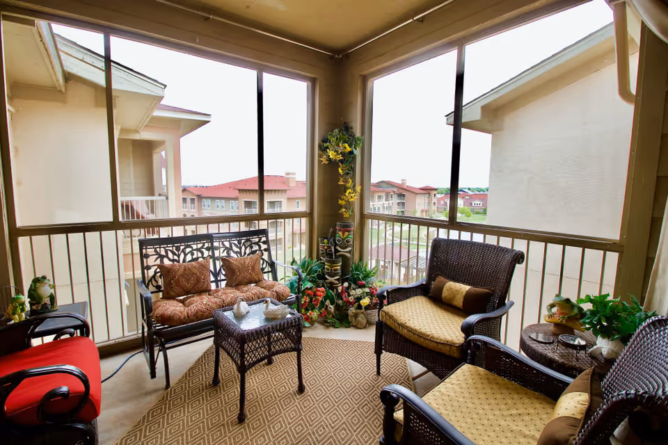 A screened-in balcony with outdoor wicker furniture including a loveseat with patterned cushions, two armchairs with yellow cushions, a small wicker table with decorative swan figurines, and various potted plants and floral arrangements. The balcony overlooks neighboring buildings with red roofs.